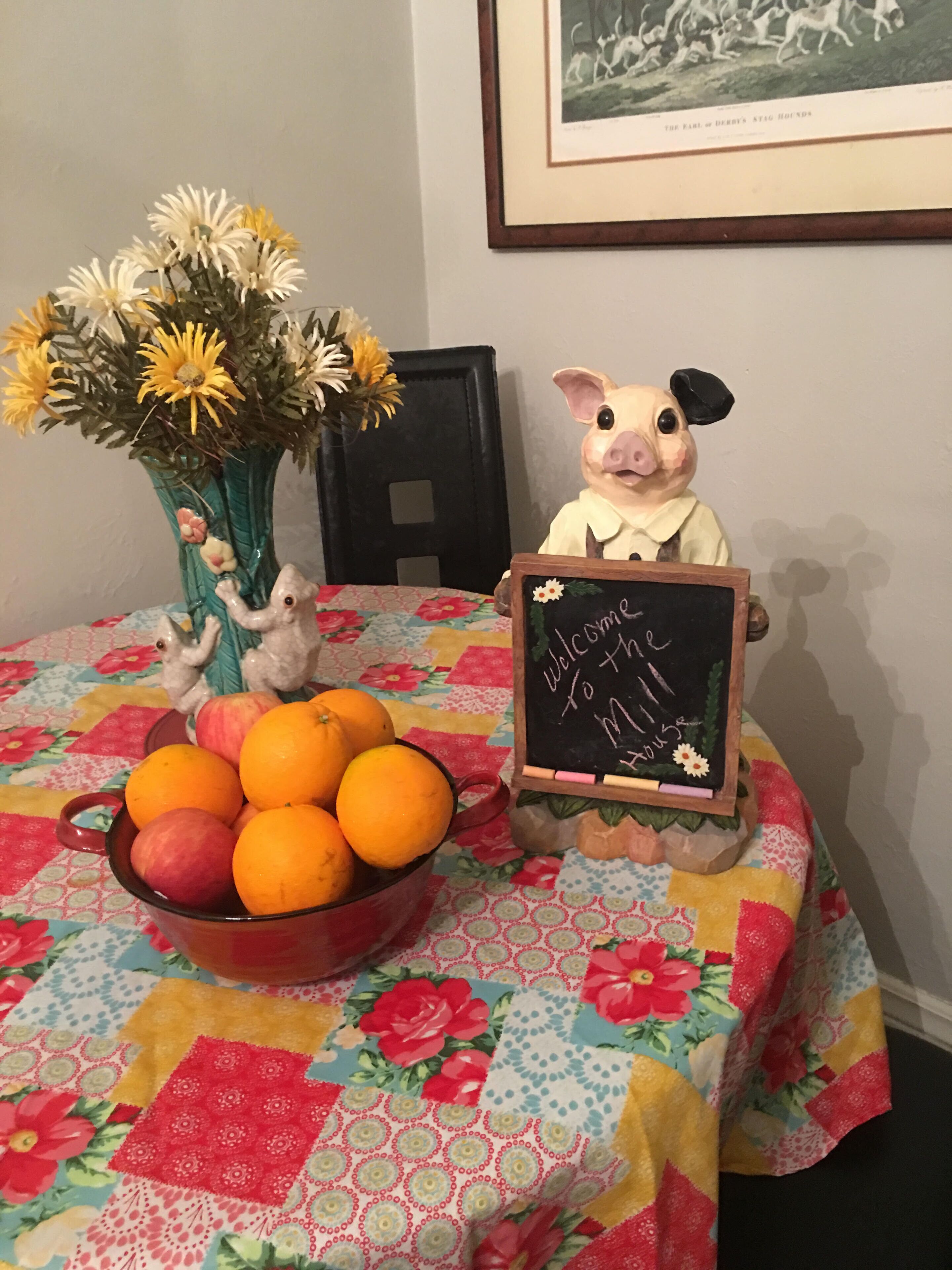 A table with a fruit bowl, a vase with flowers, and a pig figurine holding a chalkboard sign.