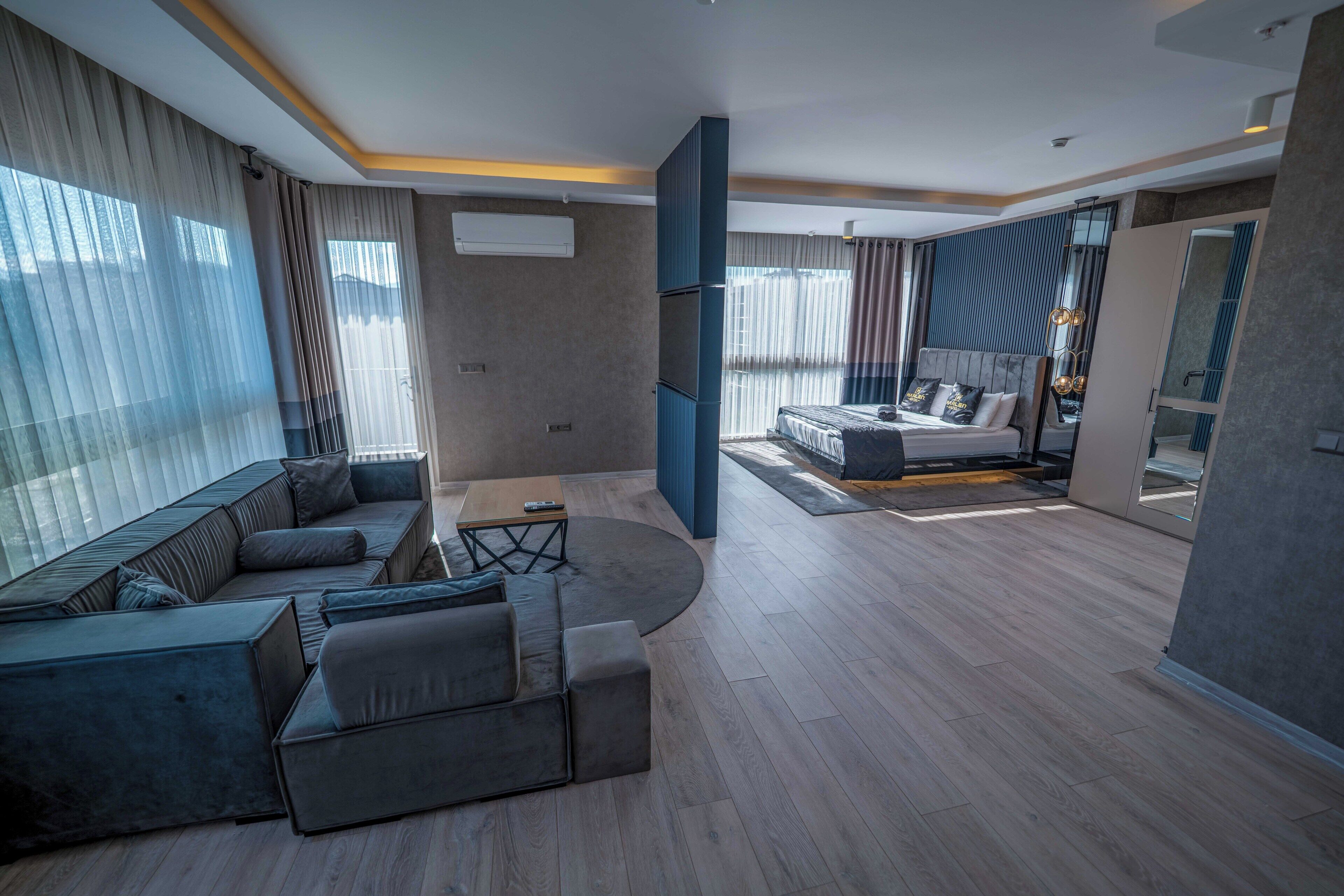 A modern hotel room with a large L-shaped sofa, a separate bed with a headboard, a small table, and a large window with curtains.