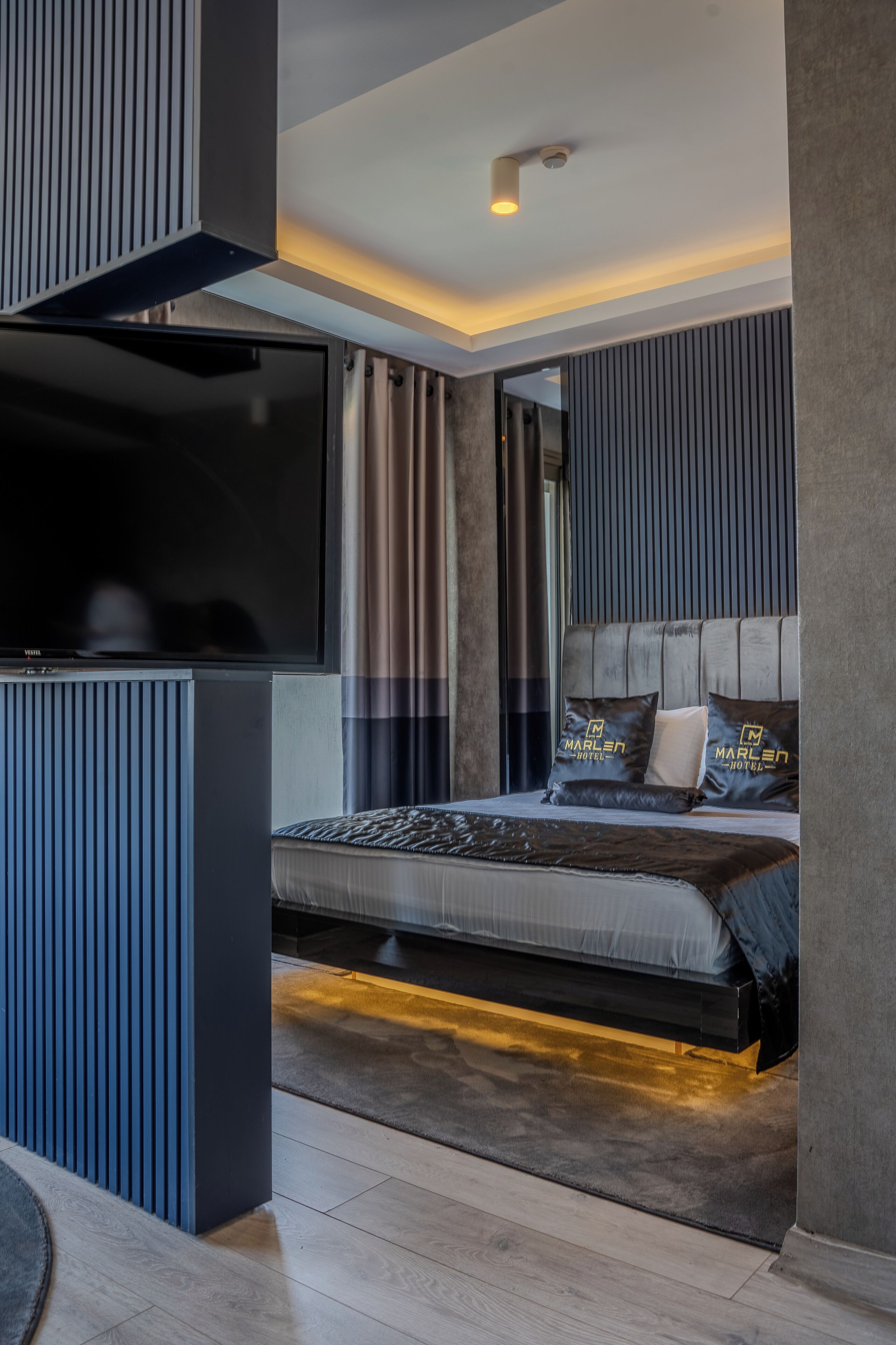 A modern hotel room with a large flat-screen TV, a bed with a headboard, and a built-in bench.