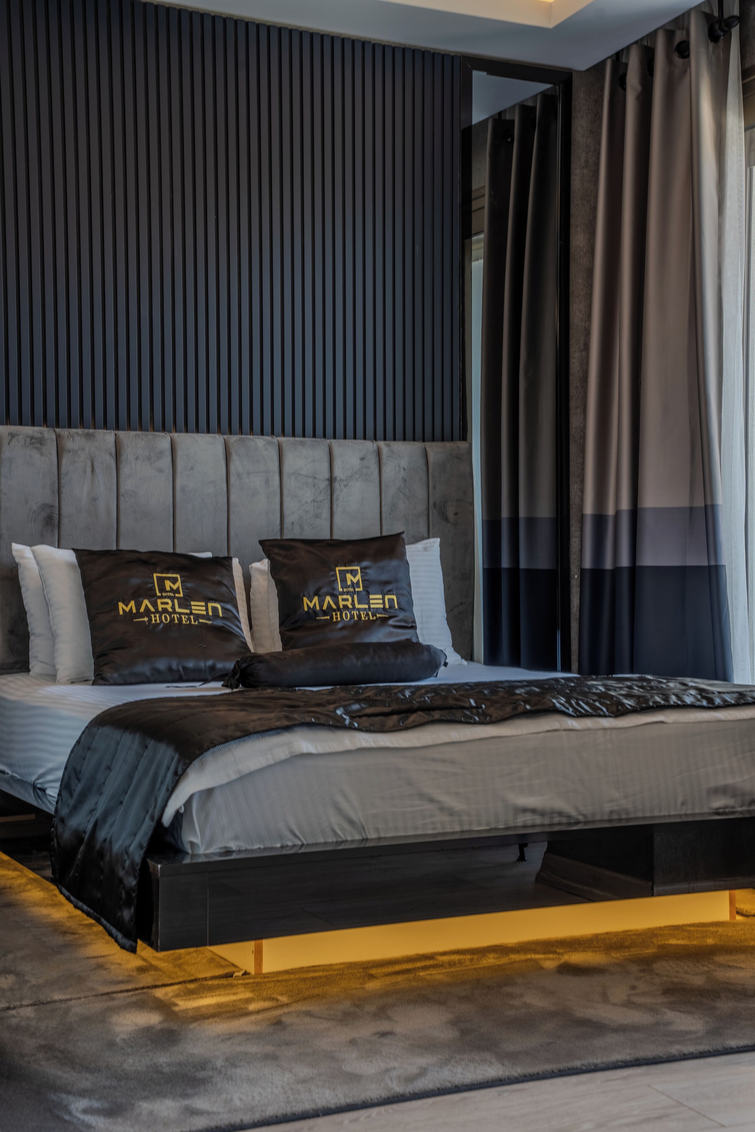 A modern hotel room with a large bed, dark headboard, and two pillows with the hotel logo.