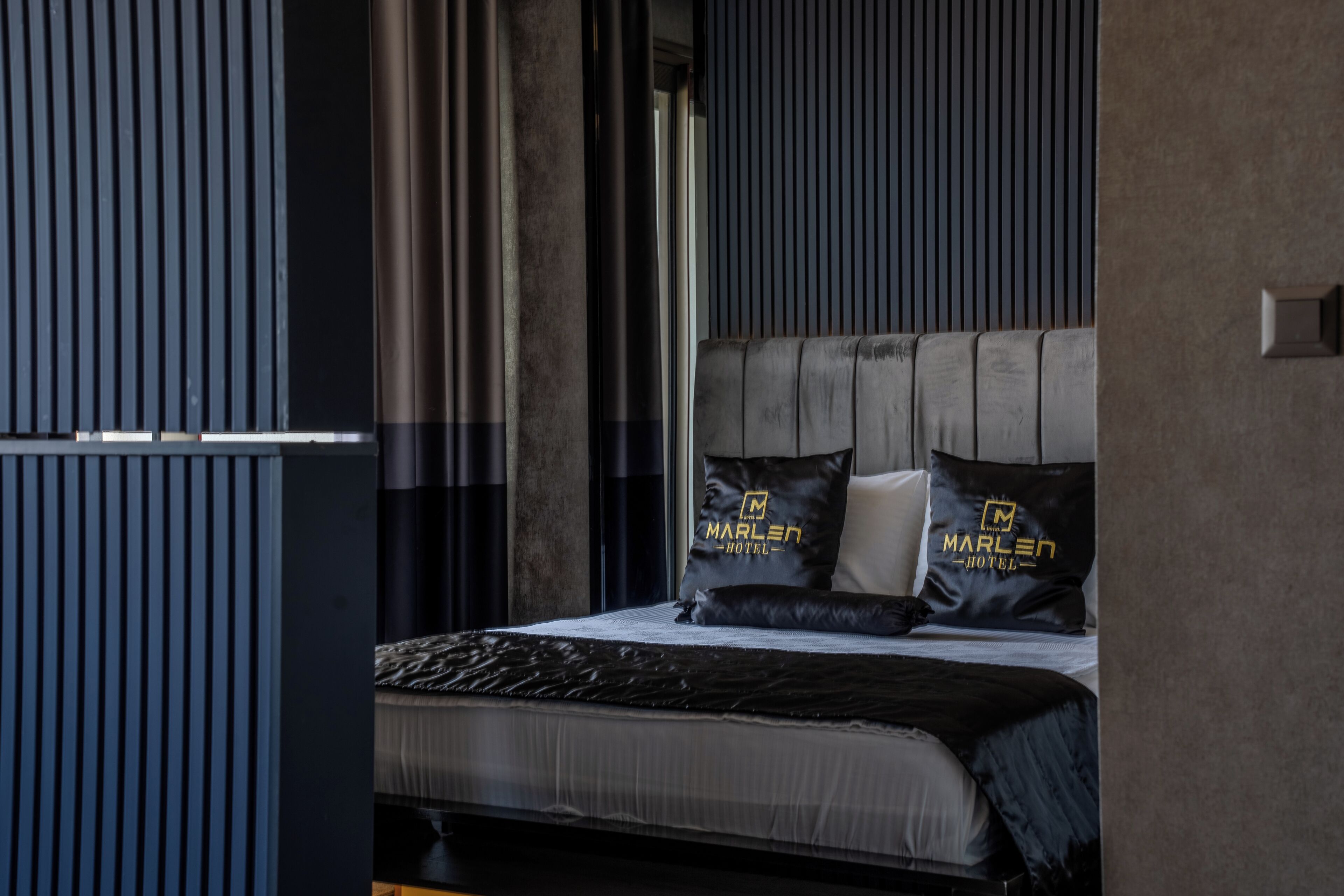 A modern hotel room with a bed, two pillows with the hotel logo, a window with blinds, and a wall with vertical lines.