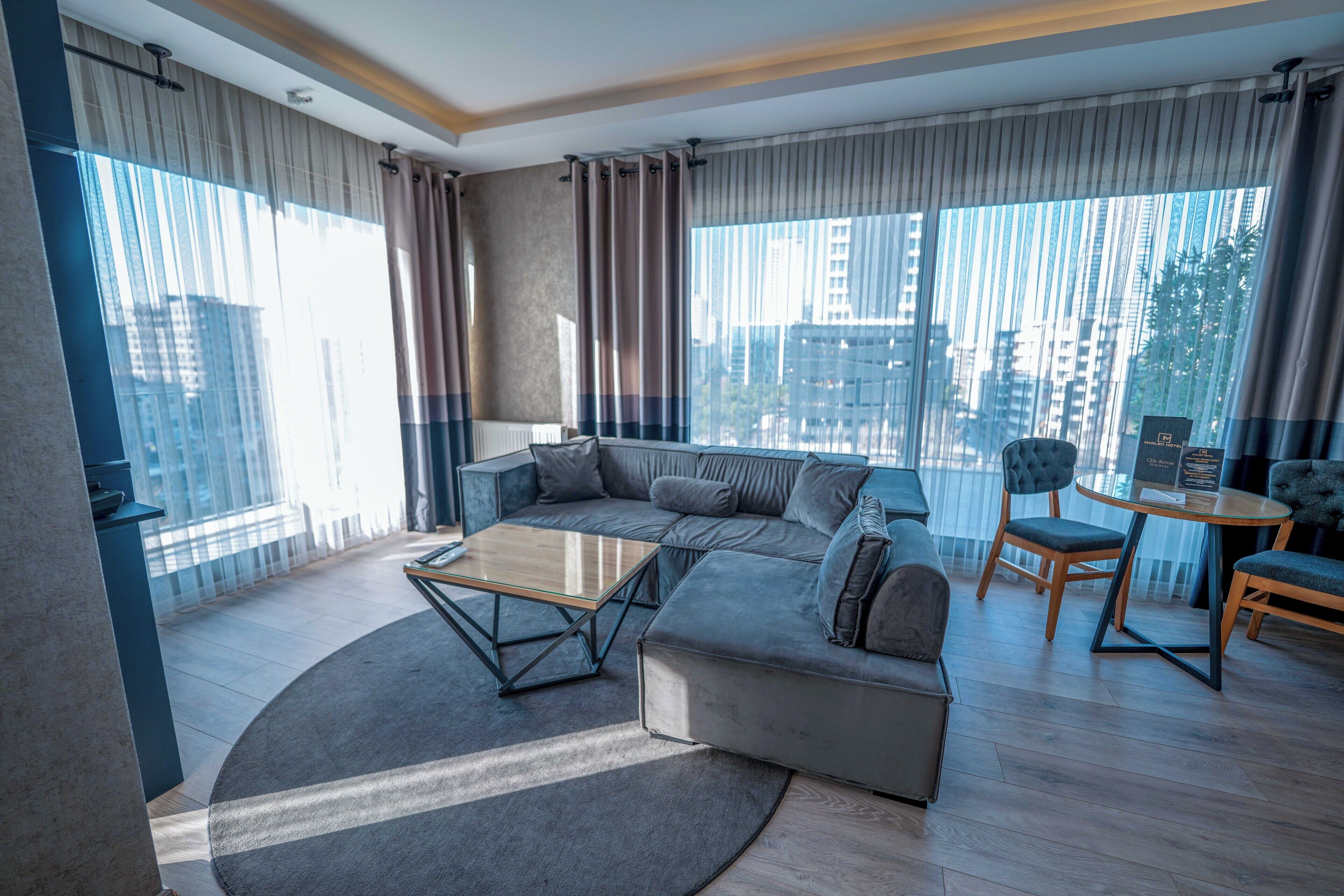A modern living room with a grey sofa, a glass coffee table, and a round rug. There are two chairs and a small round table with a menu. The room has large windows with curtains and a view of tall buildings.