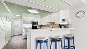 A modern kitchen with white cabinets, a central island with stools, a microwave, and a clock on the wall. - Oceanfront St Augustine Beach Condo/ Steps to Pool +  Beach (St. Augustine Beach)