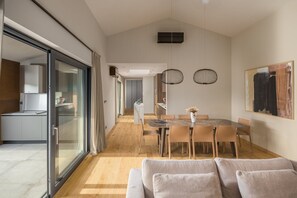 A modern dining area with a large table and chairs, a sofa in the foreground, and a kitchen visible through glass doors.
