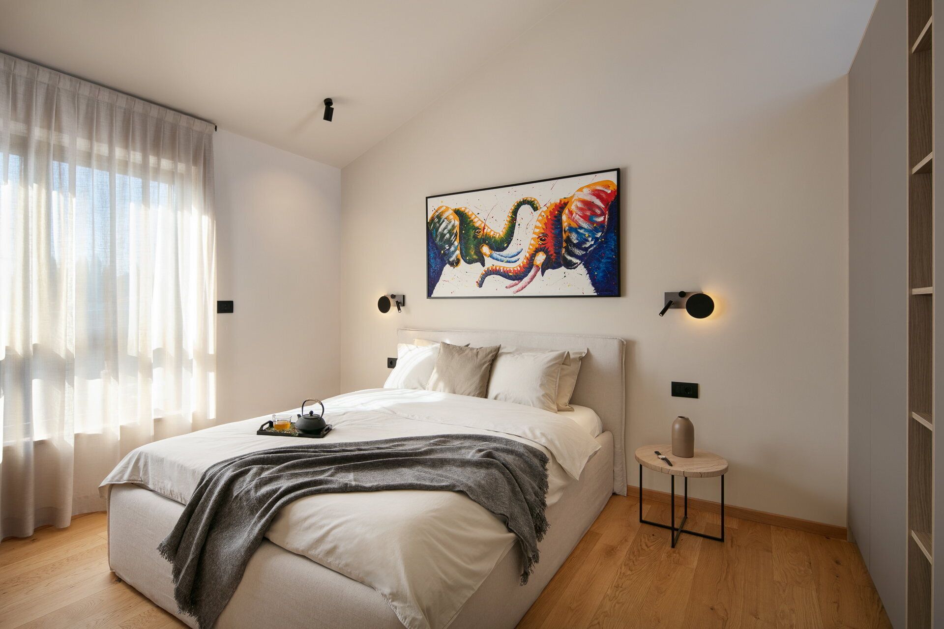 Un dormitorio moderno con una cama grande, una mesita de noche, un cuadro en la pared y una ventana con cortinas traslúcidas.