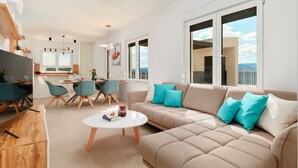 Una sala moderna con un sofá beige, sillas color turquesa y una mesa de centro blanca. Cuenta con una zona de cocina con una mesa de comedor y sillas, y amplios ventanales que ofrecen vistas al exterior.