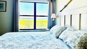 Una camera da letto con una grande finestra, un letto con lenzuola a motivi, una lampada da comodino e vista sulla spiaggia.