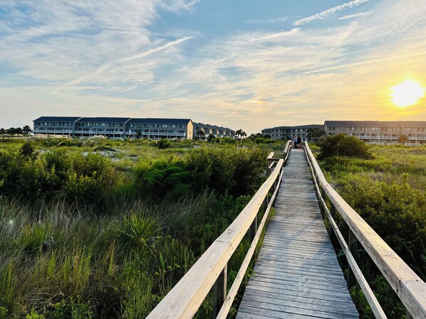 A wooden boardwalk leading to a building with multiple windows and balconies, surrounded by greenery.