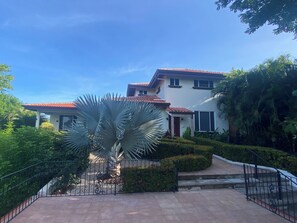 Una casa de dos pisos con techo de tejas rojas, rodeada de abundante vegetación y un jardín cuidado.