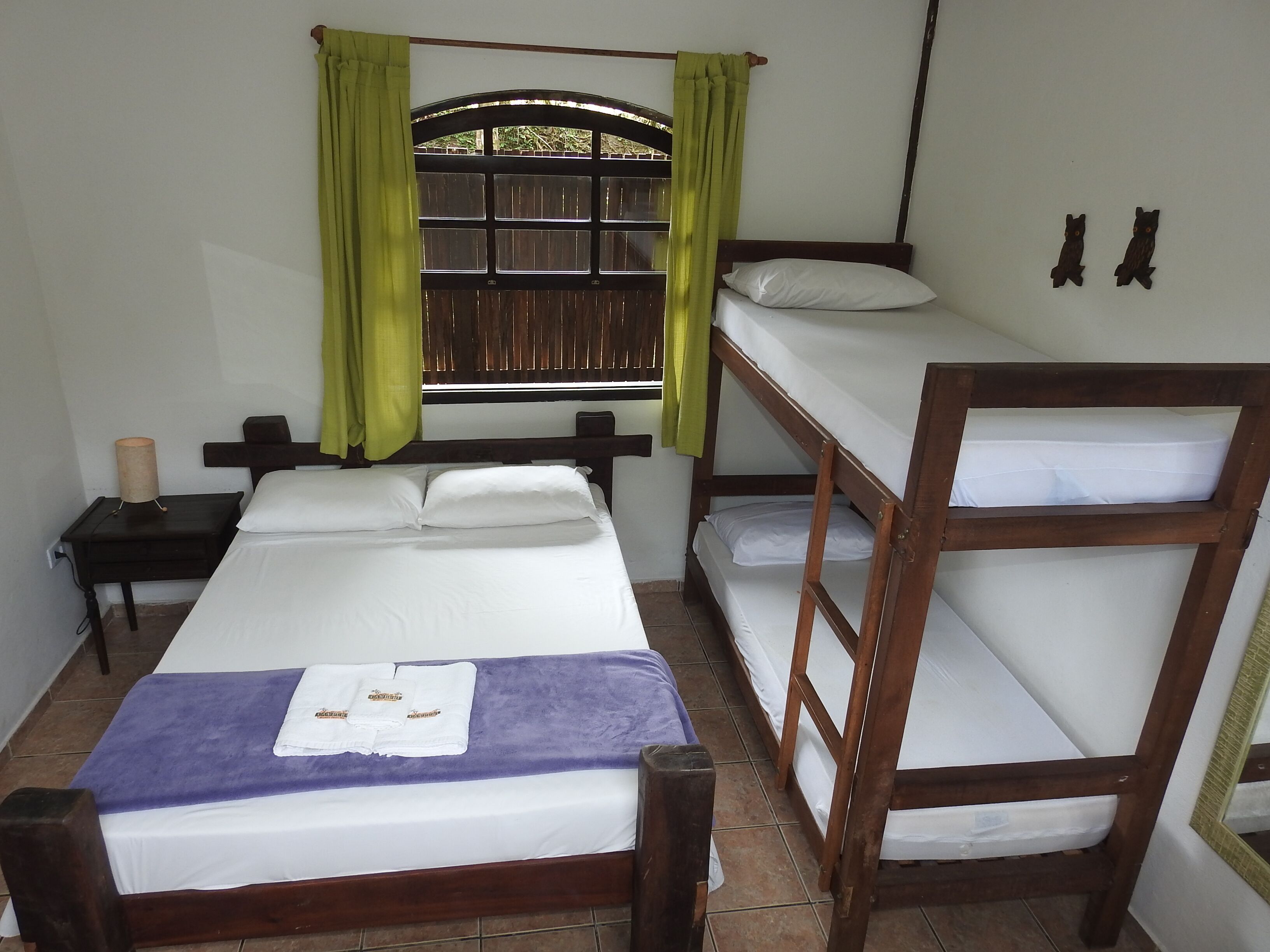 A room with a bunk bed, a single bed, a nightstand, a lamp, and a window with curtains.