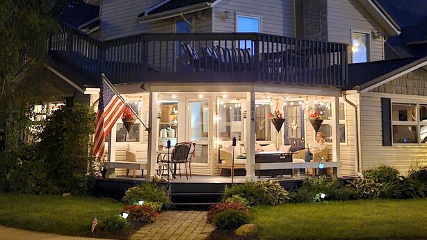 Een huis met twee verdiepingen, een overdekte veranda, een vlag en een goed verlicht interieur.