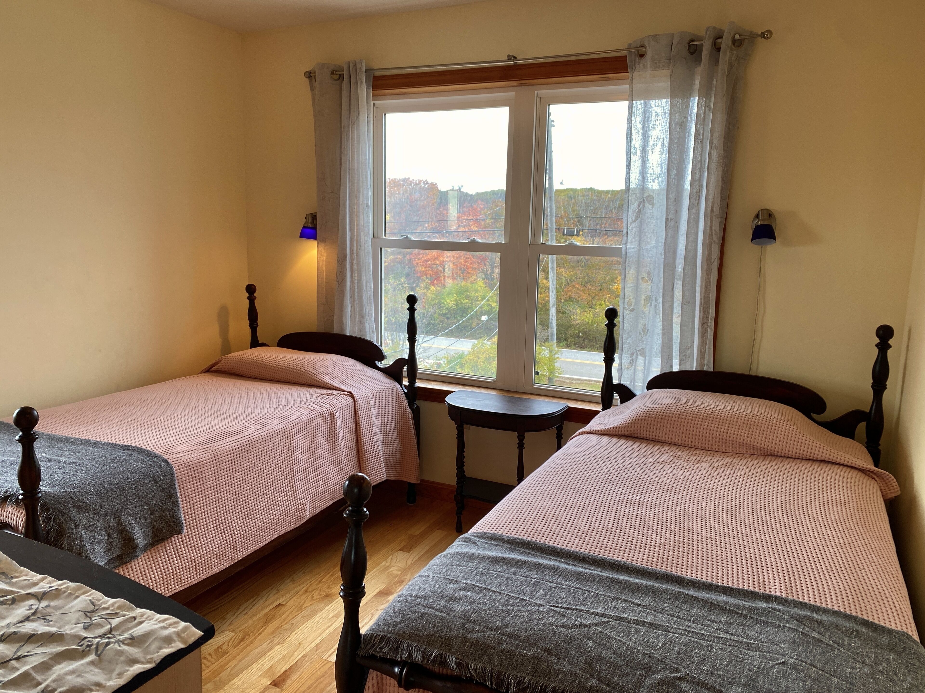 Una habitación con dos camas individuales, una mesita y un ventanal con vistas a árboles otoñales.