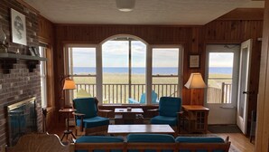 Une pièce chaleureuse donnant sur la plage, avec des murs en bois, une cheminée et des fauteuils recouverts d’un tissu bleu.