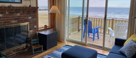 A living room with a fireplace, a large window with a view of the beach, and a deck with chairs.