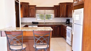 A kitchen with wooden cabinets, a granite countertop, two bar stools, a window with a view of palm trees, and a white refrigerator.