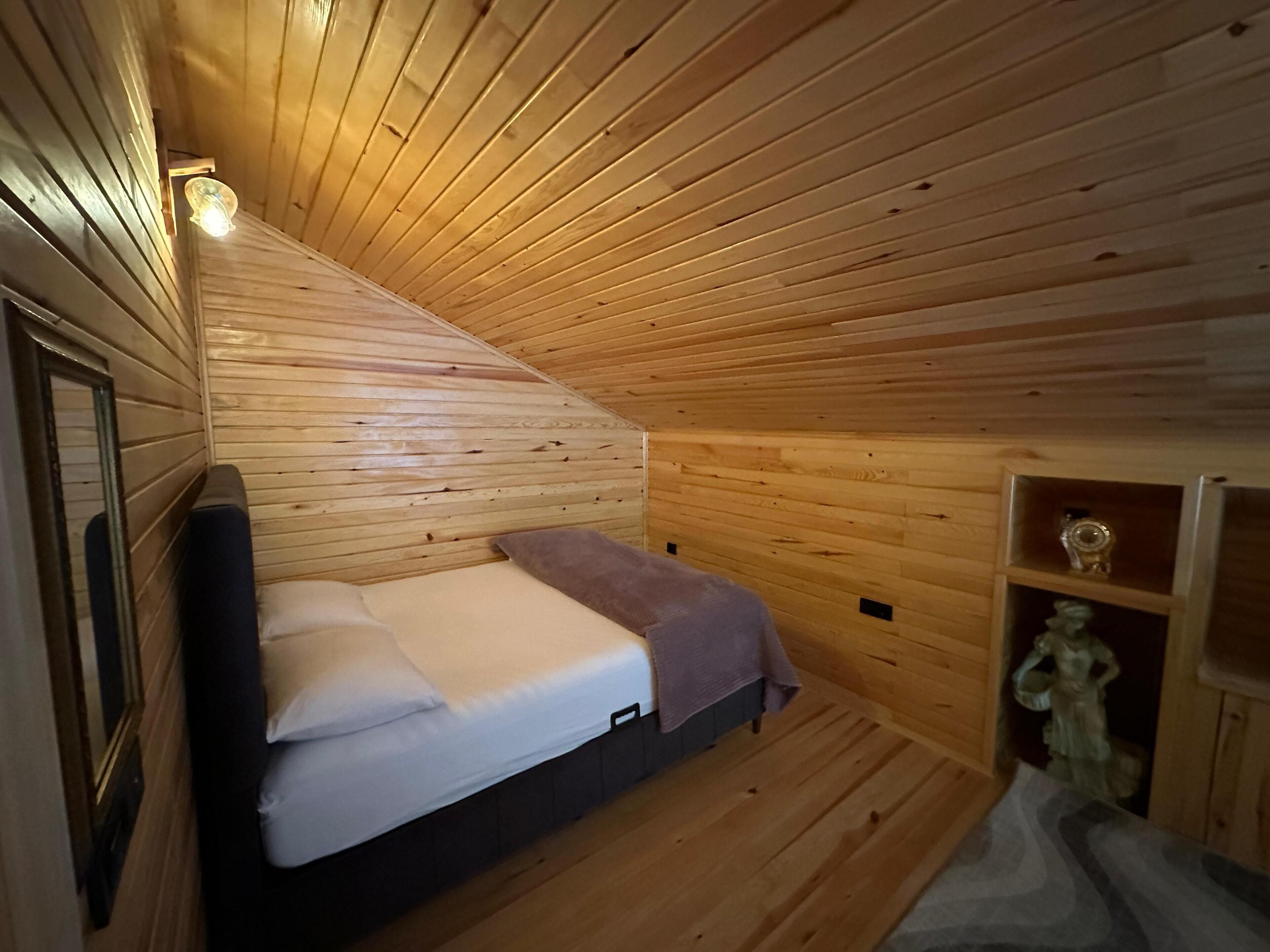 A small, cozy room with wooden walls and a sloped ceiling. It features a bed with white bedding and a purple throw blanket. There is a small shelf with decorative items and a statue.