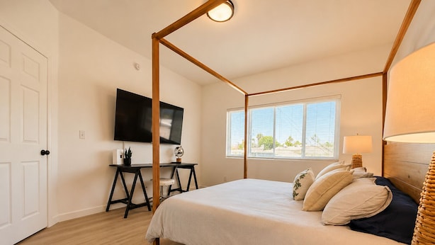 A bedroom with a four-poster bed, a desk with a chair, a television on the wall, and a window with blinds.