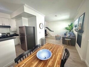 A modern kitchen with a stainless steel refrigerator, a dining table with chairs, a clock on the wall, and a staircase leading to a living area with a sofa and a plant.