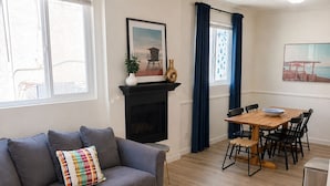 A living room with a sofa, a round coffee table, a fireplace, a dining area with a table and chairs, and a window with blinds.