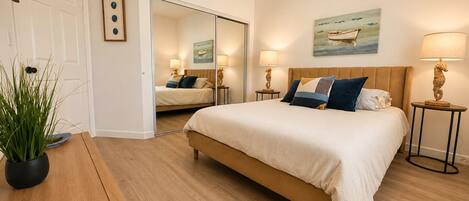 A bedroom with a bed, bedside tables, a mirror, a potted plant, and a painting on the wall.
