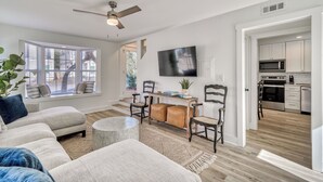 A spacious living room with a large sectional sofa, a coffee table, and a TV mounted on the wall. There is a dining area with a wooden table and chairs, and a kitchen visible through an open doorway. - Golf Cart & Pool included! Pet-Friendly Retreat <1 mile from Beach (Santa Rosa Beach)