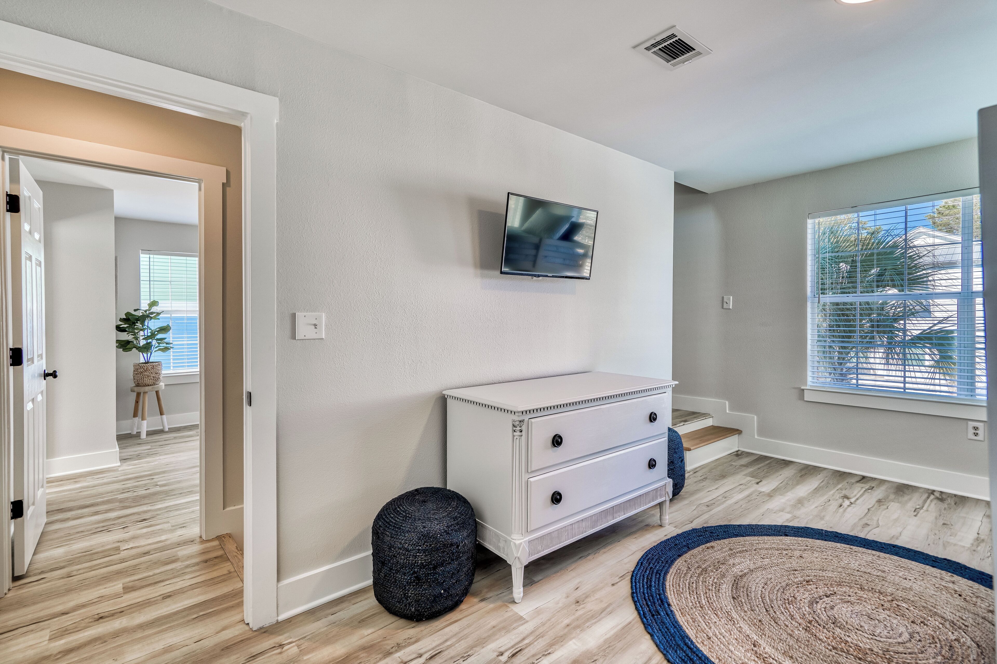 A room with a white dresser, a television mounted on the wall, a black ottoman, and a blue and beige rug.