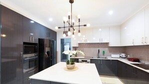 A modern kitchen with dark cabinets, stainless steel appliances, a central island with a vase of flowers, and a unique chandelier. - Zenhouse Stylish: Lawn ~ Workspace ~ OutdoorDining (Muirhead)