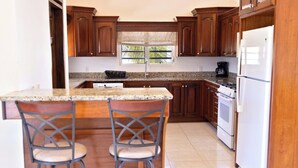 A kitchen with wooden cabinets, a granite countertop, two bar stools, a window with a view of palm trees, and a white refrigerator. - Sienna is Private, Tranquil,  and Perfectly Located near Shoal Bay Beach! (Shoal Bay)