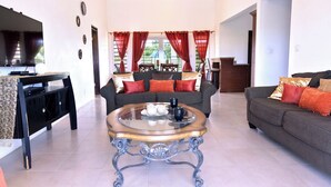 A spacious living room with a glass coffee table, two sofas with red cushions, a TV, and a dining area with red curtains. - Sienna is Private, Tranquil,  and Perfectly Located near Shoal Bay Beach! (Shoal Bay)