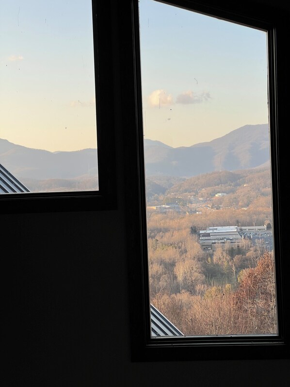 Vista a través de una ventana, que incluye un edificio de techo plano y un paisaje con árboles y montañas a lo lejos.