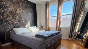 Een moderne slaapkamer met een groot bed, een decoratief behang, een groot raam met gordijnen en een spiegel.