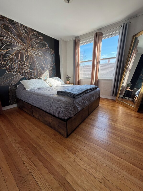 A modern bedroom with a large bed, a decorative wall mural, a large window with curtains, and a mirror.
