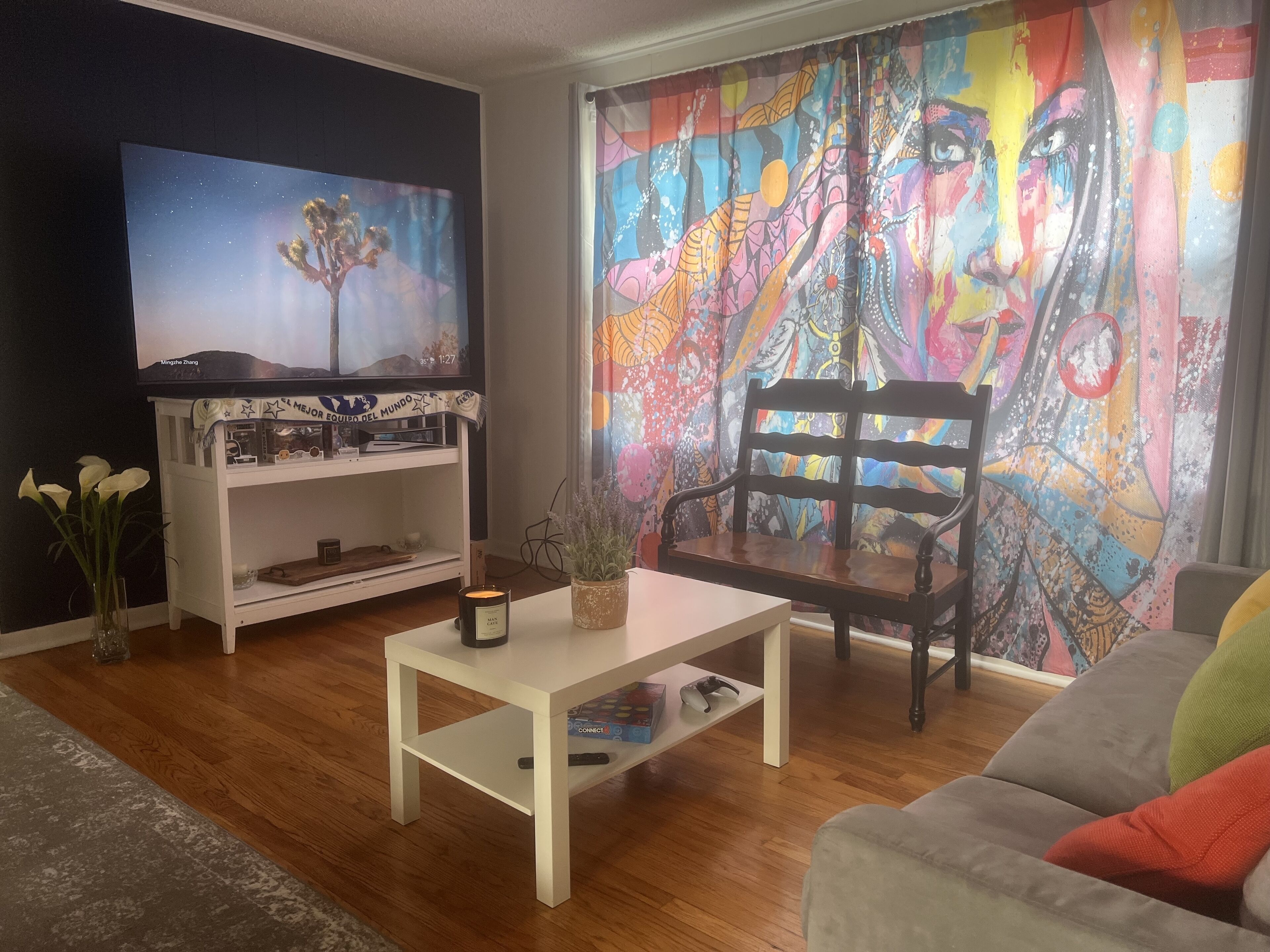 Een woonkamer met een televisie, een witte salontafel, een houten stoel en een kleurrijk abstract schilderij aan de muur.