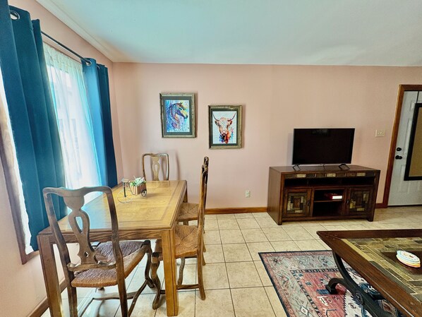 A dining area with a wooden table and chairs, a television on a cabinet, and two framed artworks on the wall.