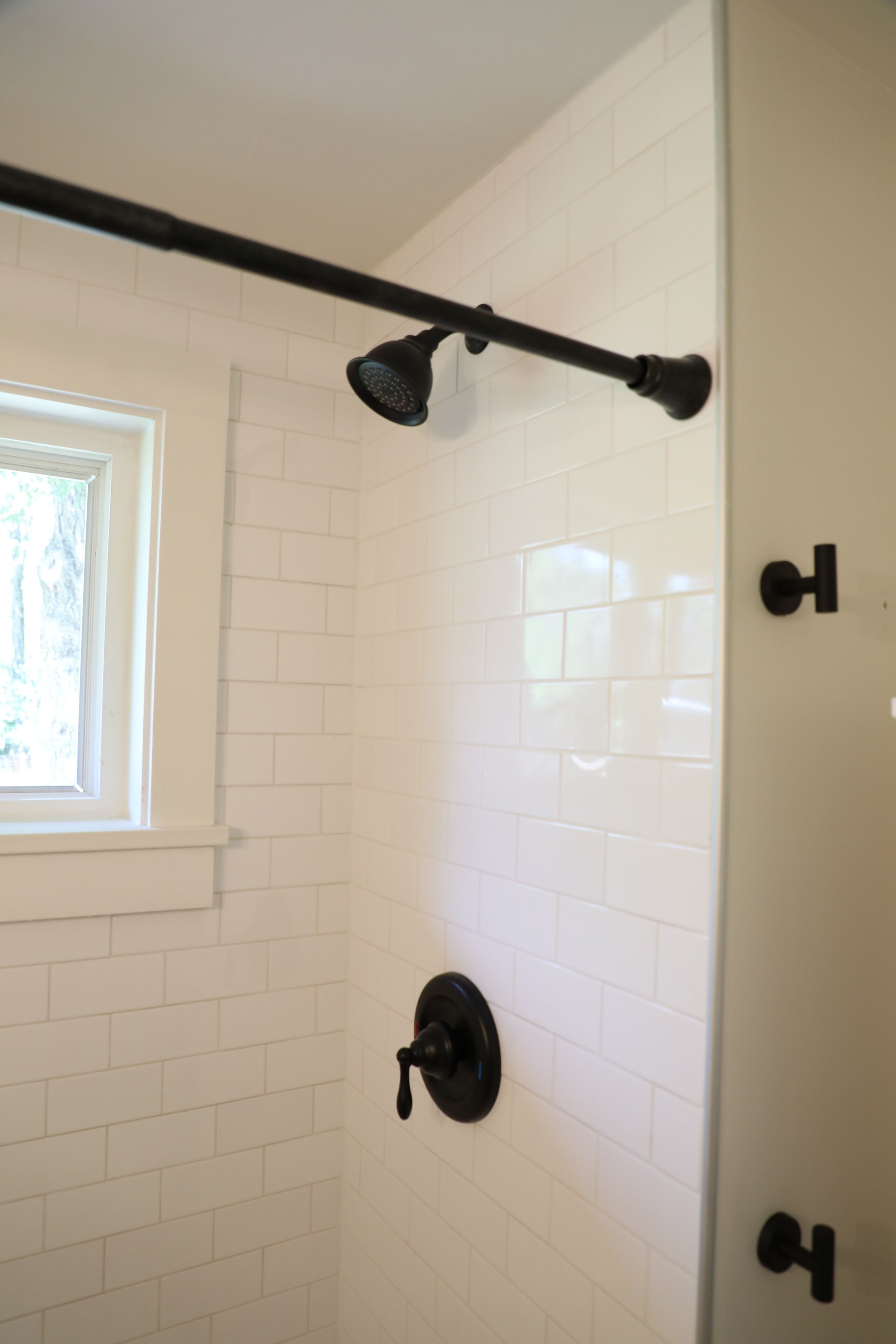 Une douche aux murs recouverts de carreaux blancs, avec une pomme de douche noire et une fenêtre à cadre blanc.
