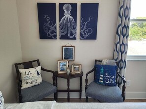 Ein Zimmer mit zwei Holzstühlen, jeder mit einem Kissen mit inspirierenden Zitaten. Ein kleiner Tisch zwischen den Stühlen trägt gerahmte Bilder und eine kleine Dekoration. Darüber hängen drei dunkelblaue Wandbehänge mit weißen Oktopusmotiven.