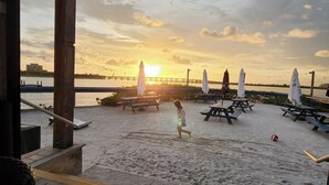 Puinen terassi, jolta avautuu näkymä hiekkarannalle. Terassilla on piknikpöytiä ja aurinkovarjoja, ja taustalla näkyy auringonlasku.