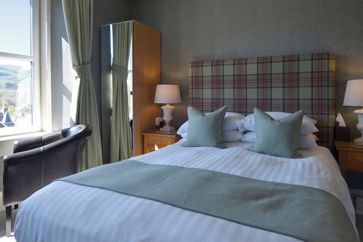 A hotel room with a bed, a chair, a nightstand with a lamp, and a plaid headboard.