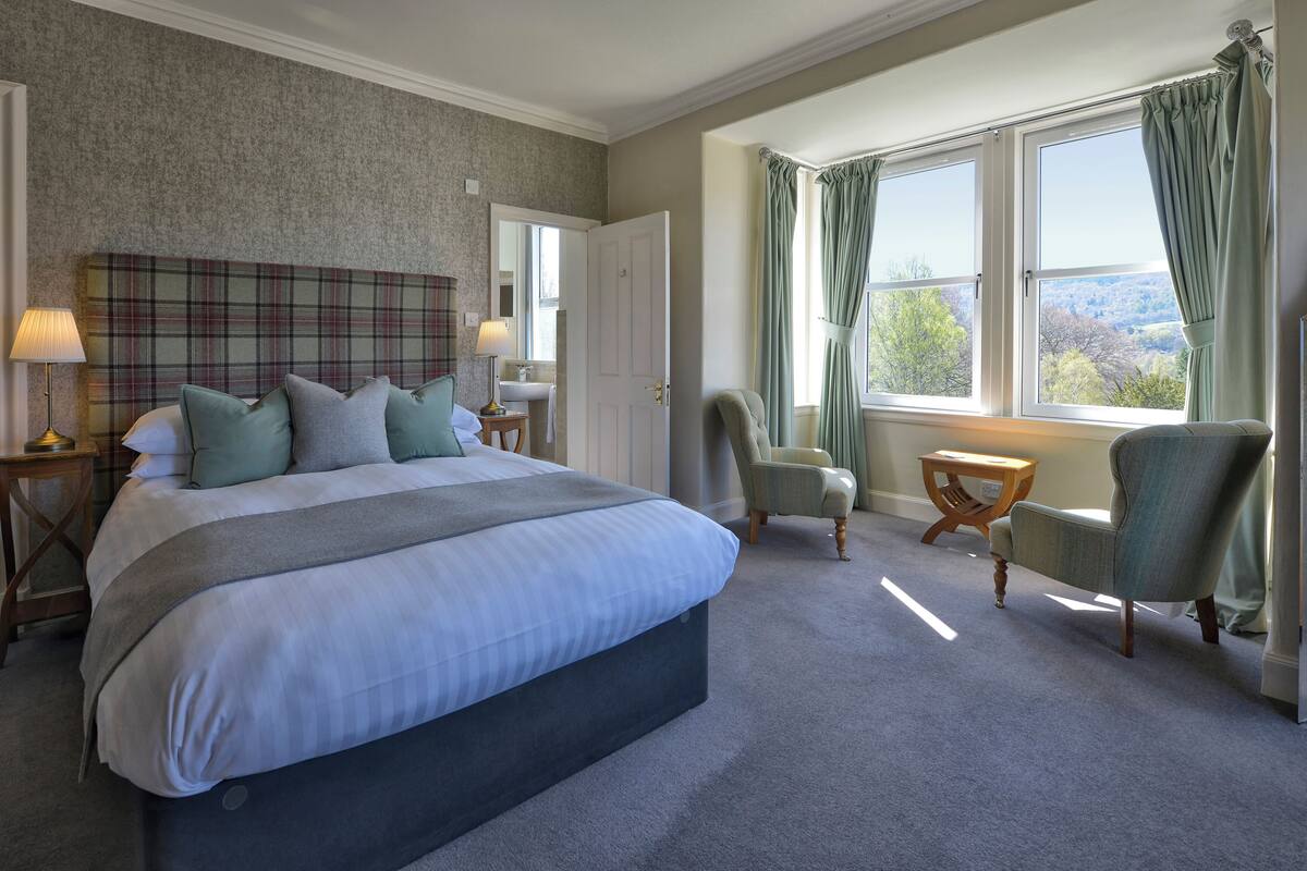 A bedroom with a large bed, a bedside table with a lamp, a chair, a small table, and a window with curtains.