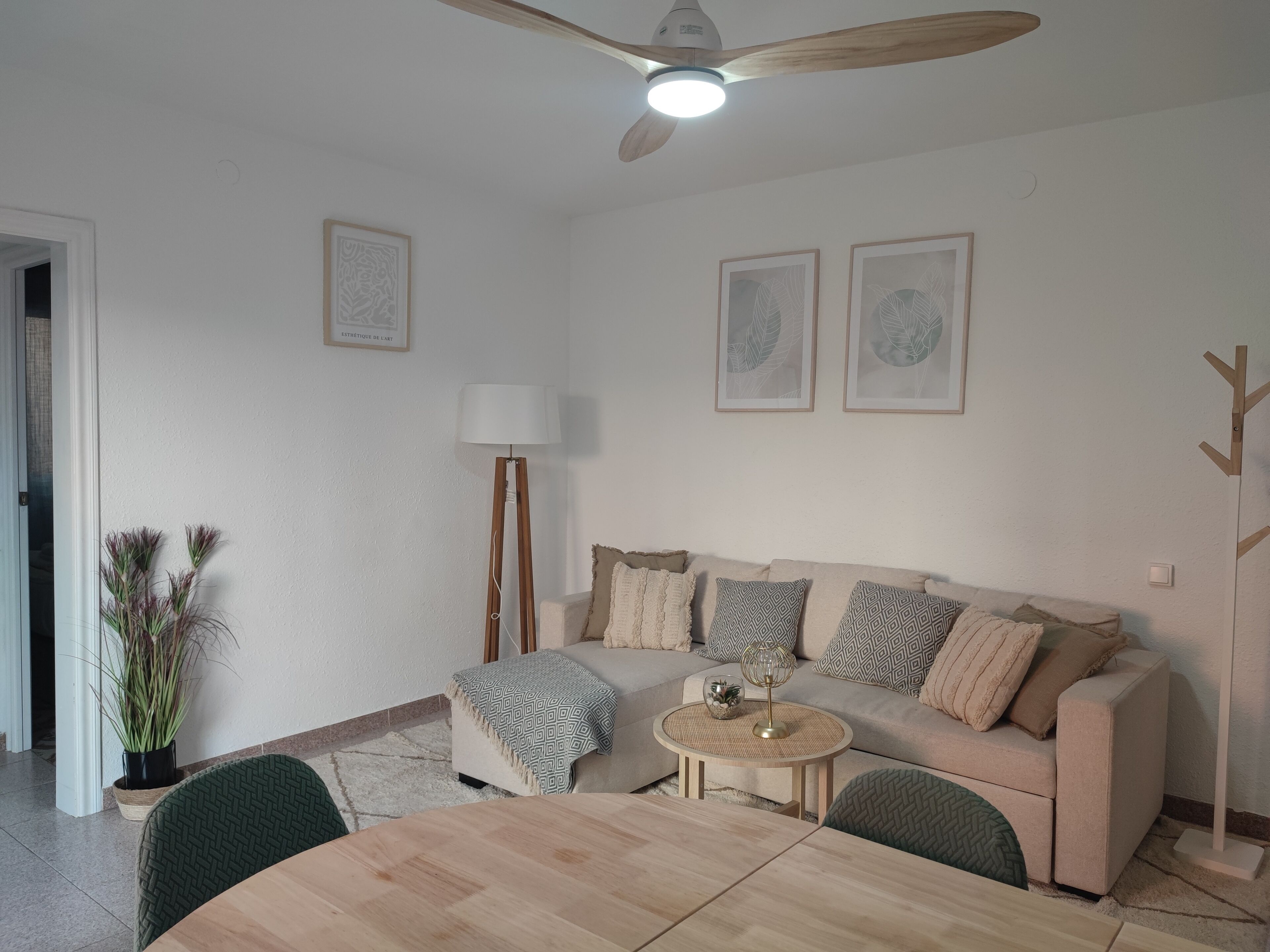 A living room with a round wooden table, a beige sofa with patterned cushions, a floor lamp, and two framed wall art pieces.