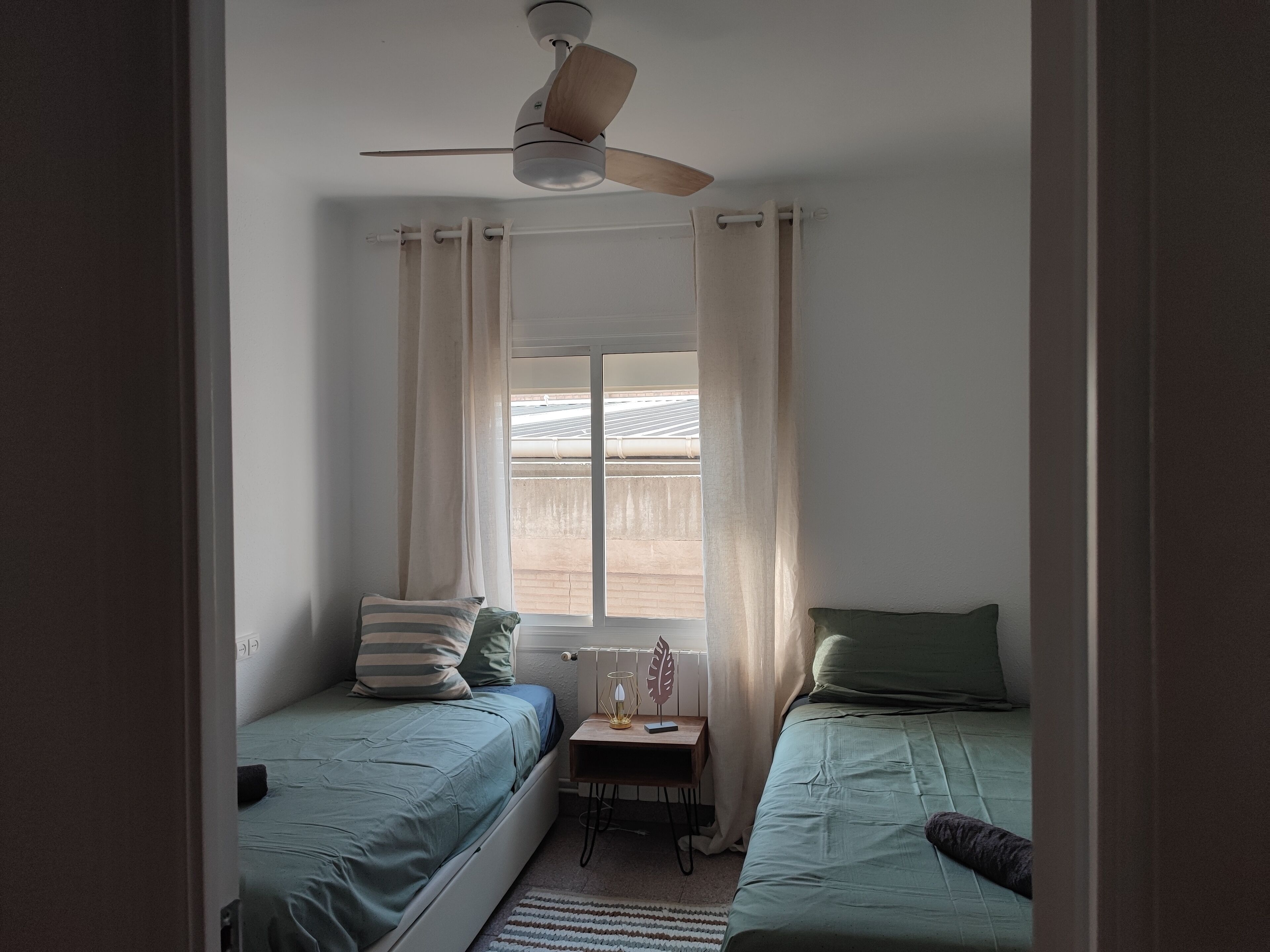A bedroom with two single beds, a ceiling fan, a window with curtains, and a small table with a lamp.