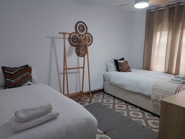 A hotel room with two beds, a wooden drying rack, patterned rugs, and a window with curtains.