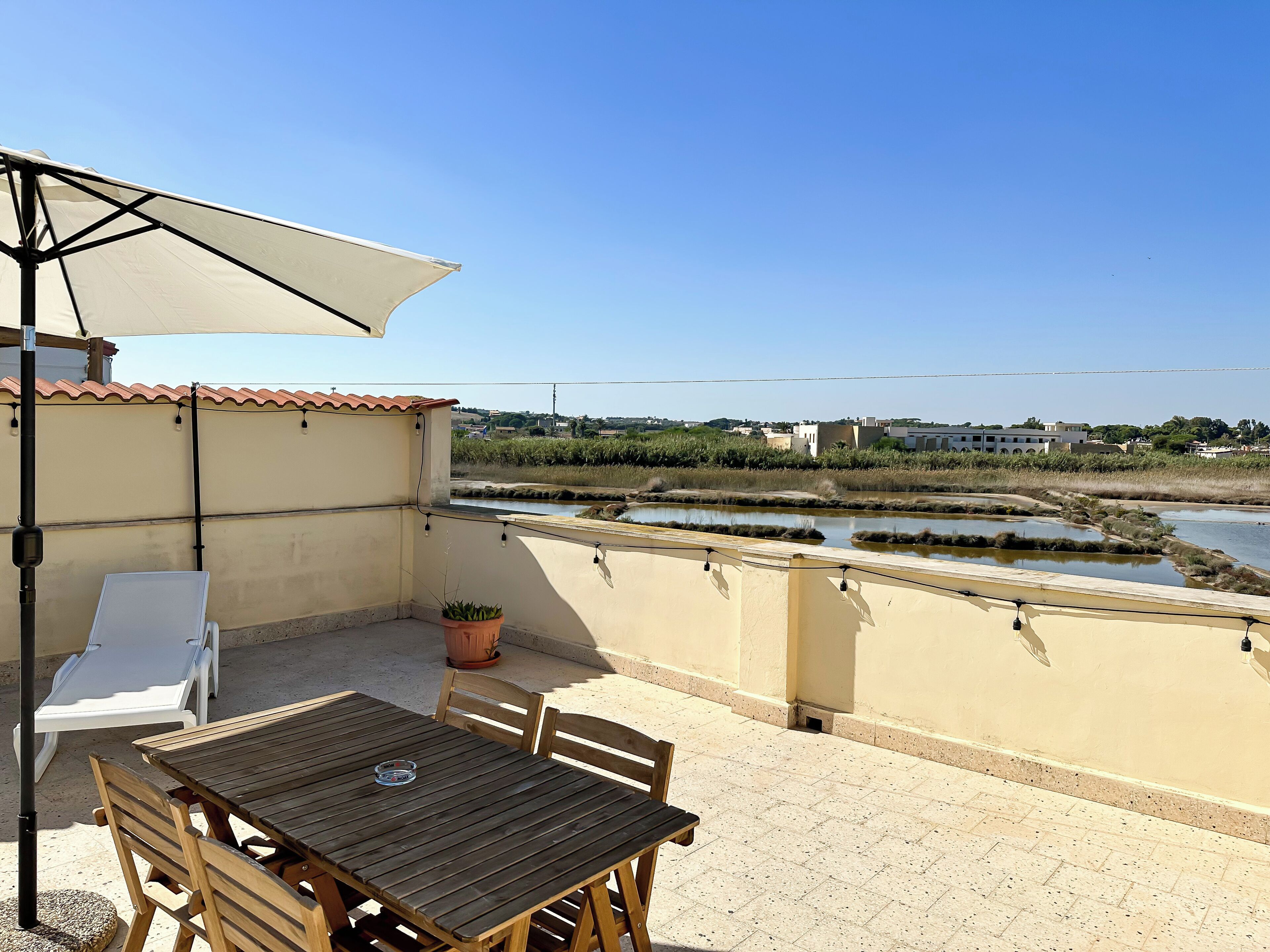 A balcony with a table and chairs, a potted plant, and a view of a water body and buildings in the distance.