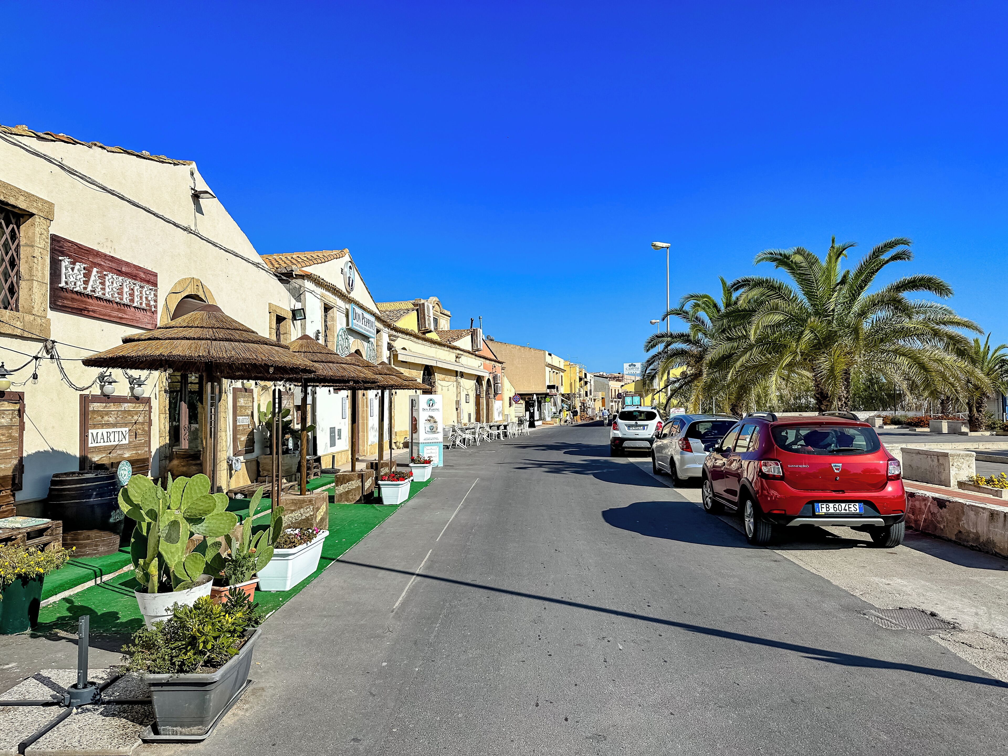 Una calle con coches aparcados, un café con terraza y palmeras.