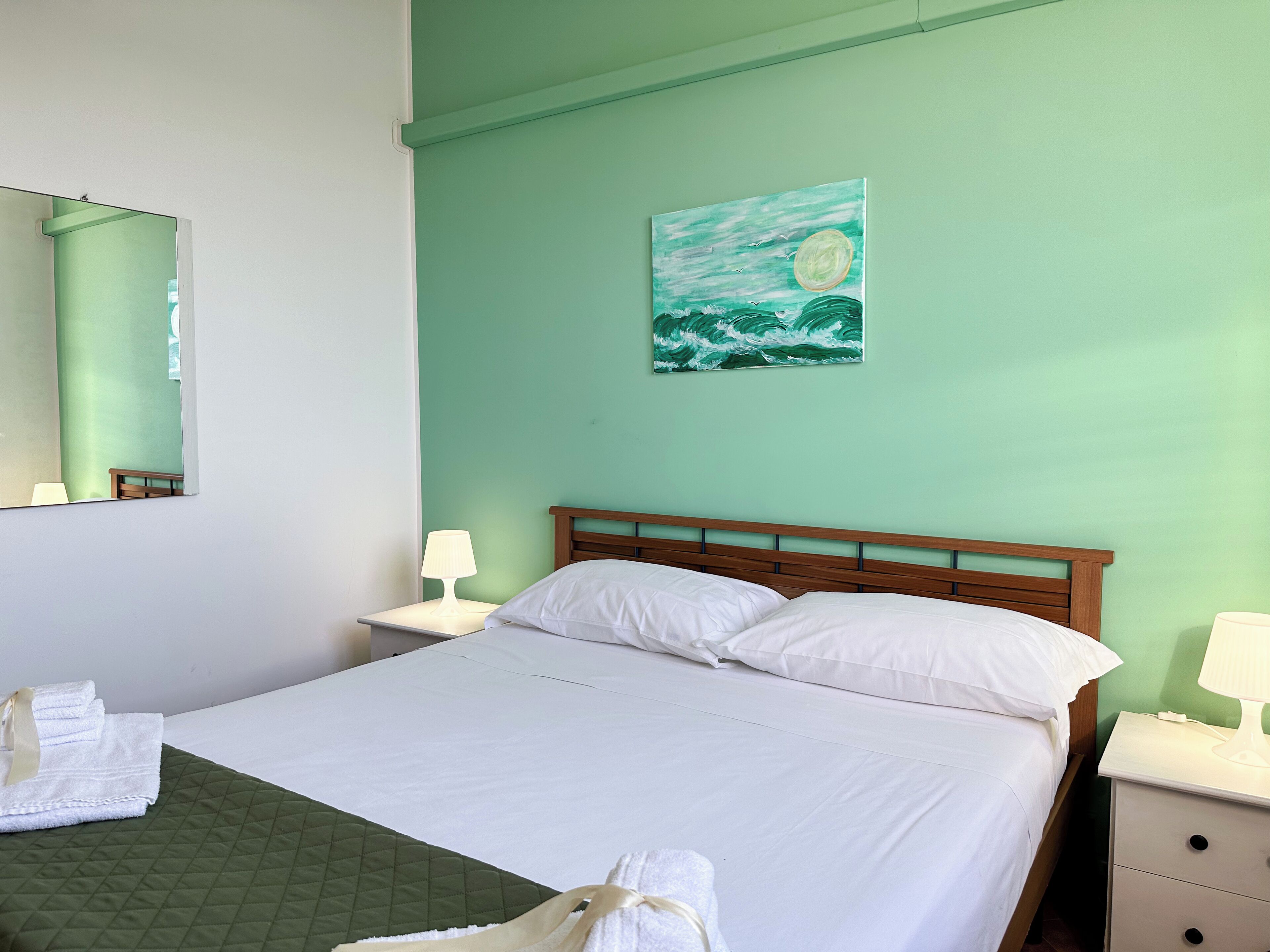 A neatly made bed with white linens, a green quilted bedspread, and a painting of waves on the wall.