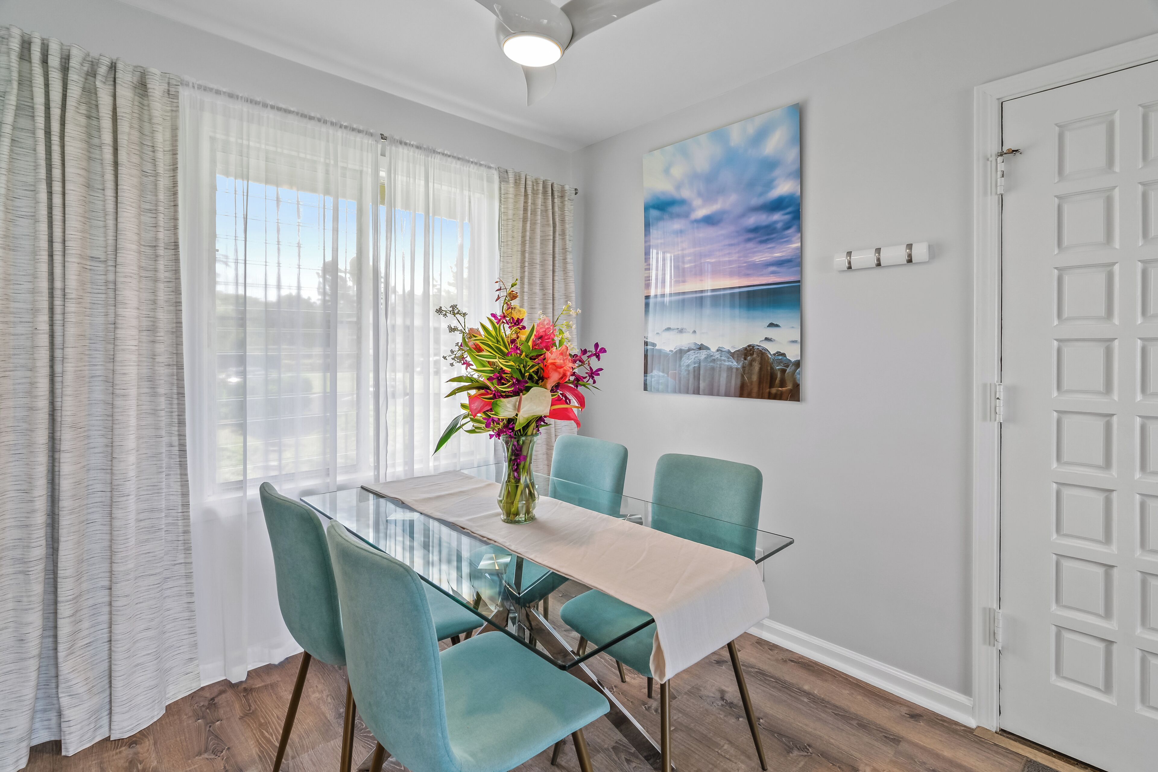 A dining area with a glass table, teal chairs, and a vase with flowers.