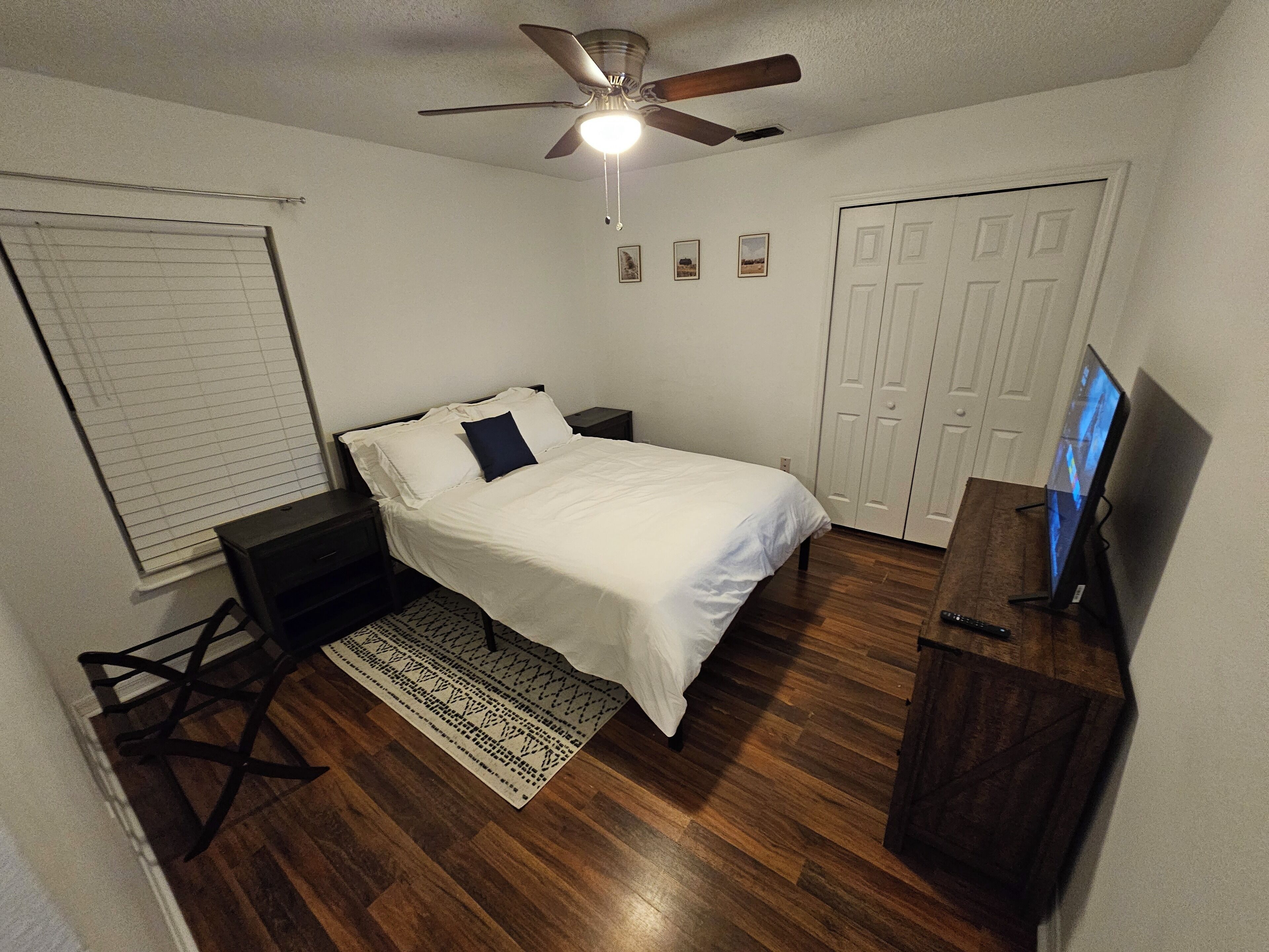 A bedroom with a bed, a ceiling fan, a TV, a wardrobe, and a window with blinds.