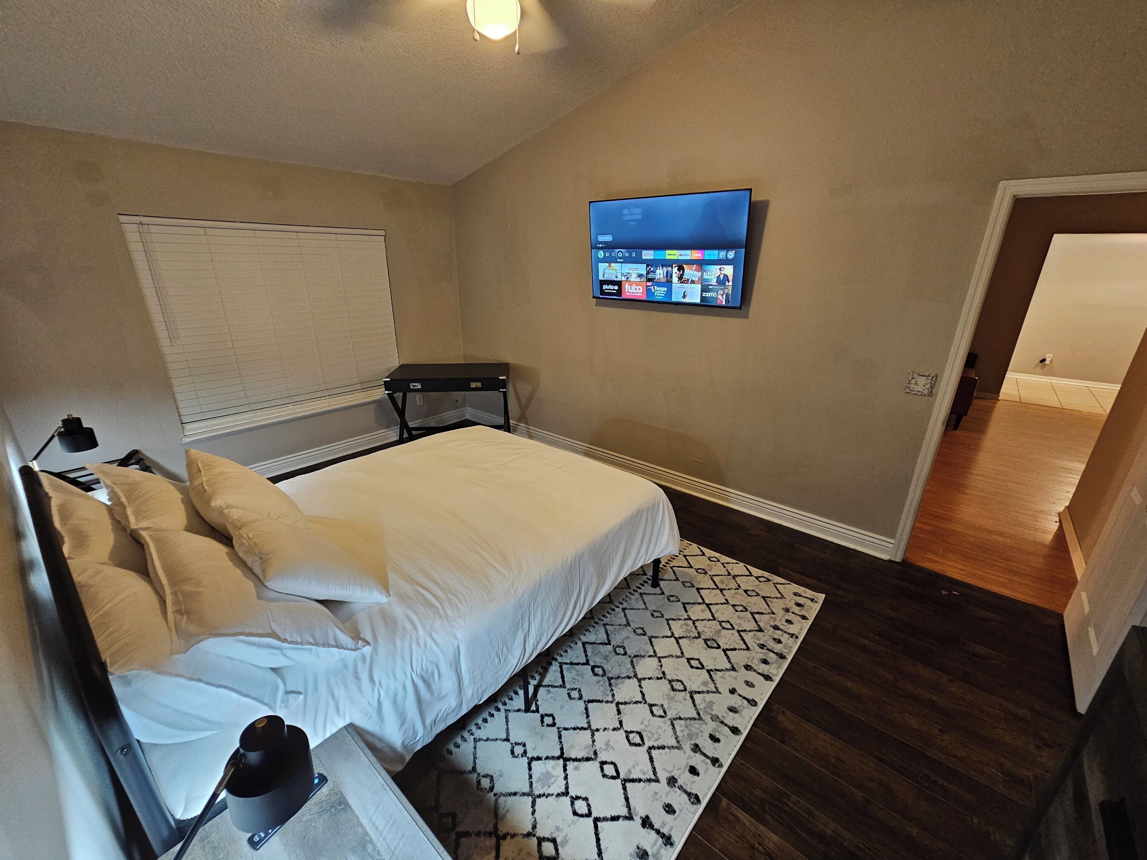A bedroom with a bed, a TV mounted on the wall, a bedside table, and a patterned rug.