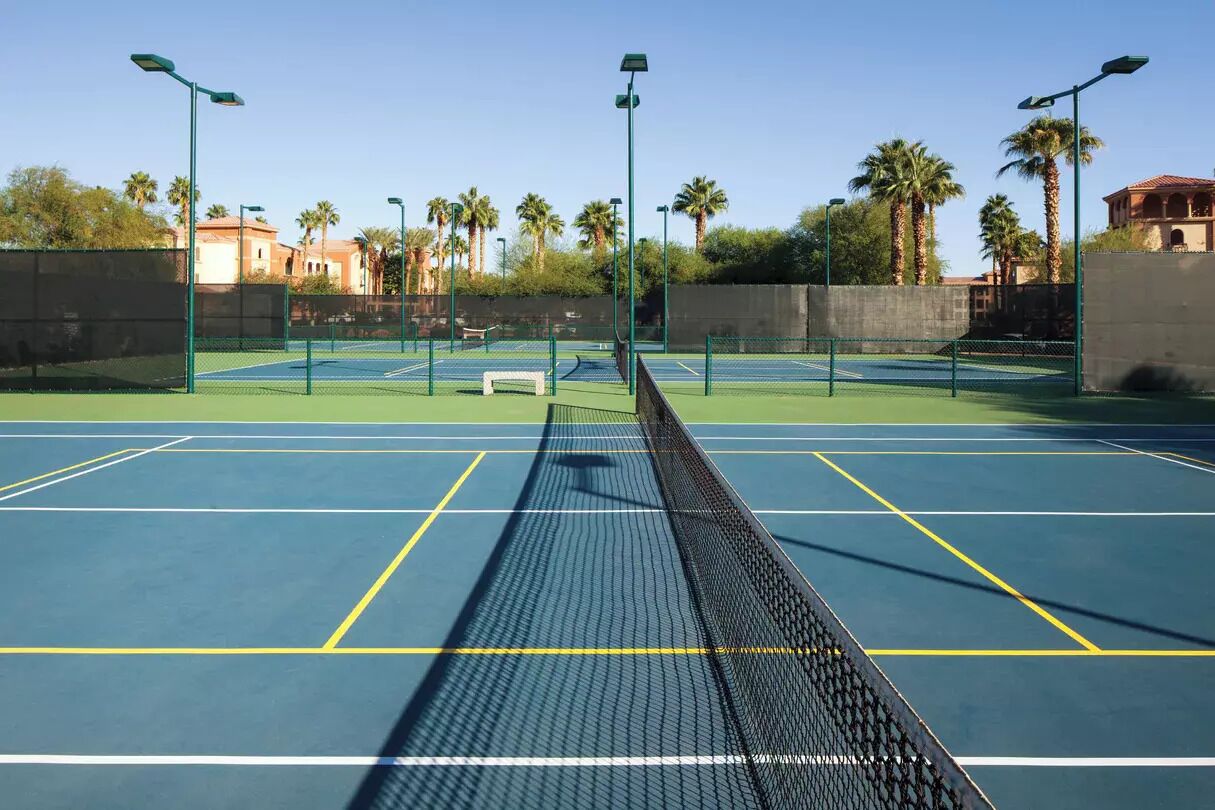 Ein Tennisplatz im Freien mit Netz, umgeben von Palmen und einem Zaun.