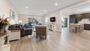 Una sala moderna con sofá, sillón y mesa de comedor. Un televisor está montado en la pared, y se observa un área de cocina.